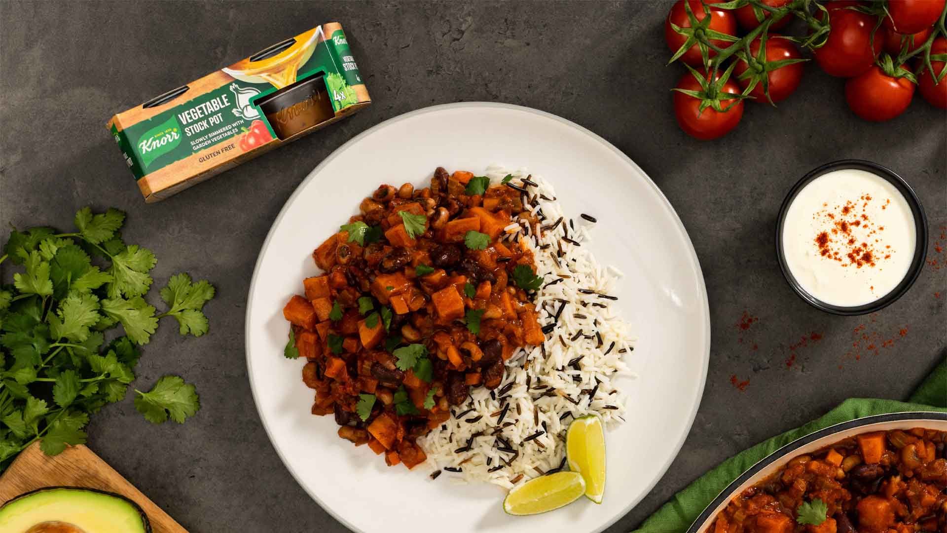 A plate of vegetable curry with rice and lime on a grey surface, encircled by a stock pots pack, parsley, dip, and tomatoes