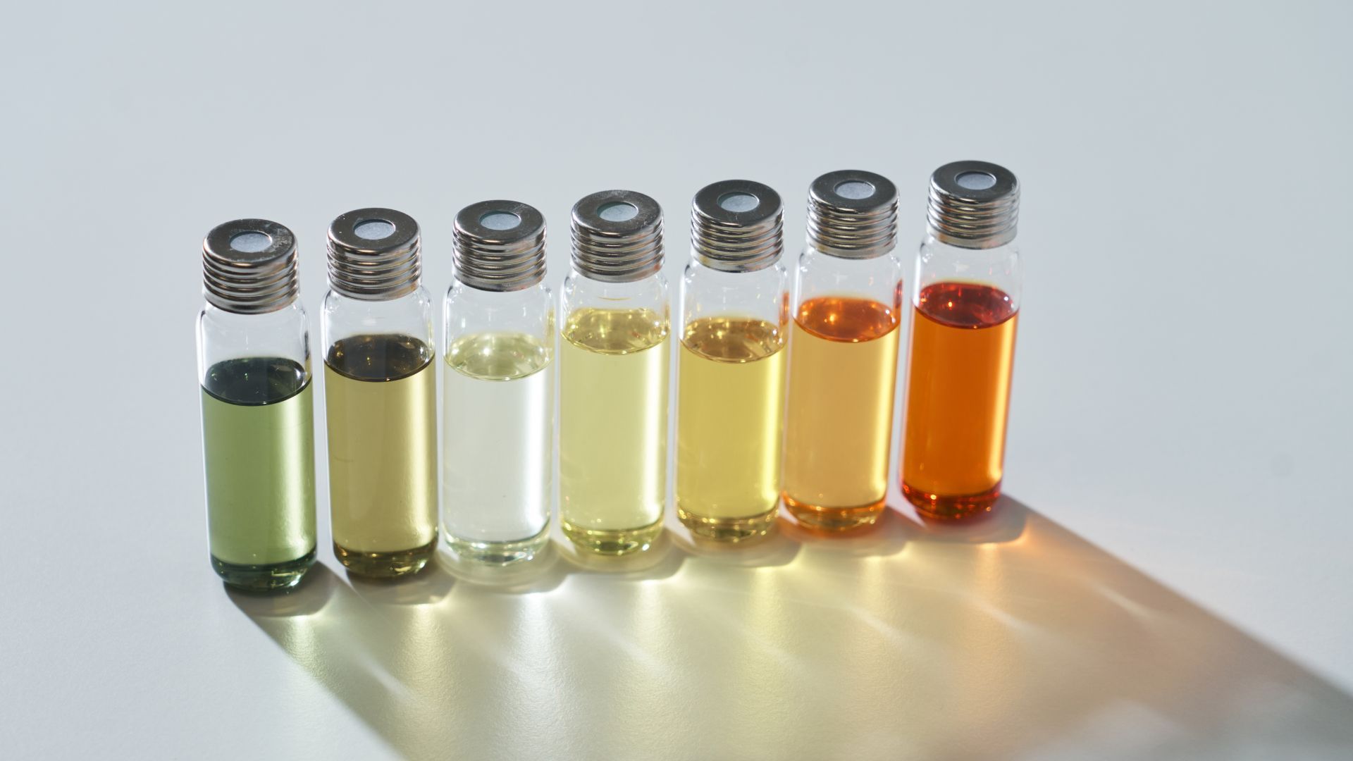 A row of seven glass vials with silver caps. They are filled with green, yellow, amber and colourless perfume samples.