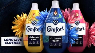 Bottles of Comfort fabric conditioner in yellow, blue and pink lined up. The background is dark denim material. Text reads: Long live clothes.
