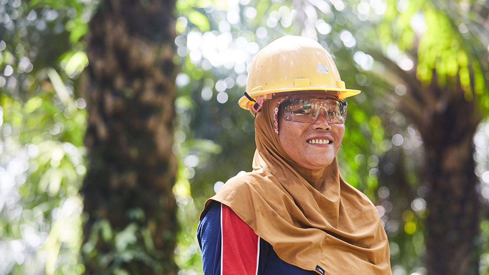 A woman in a headscarf and yellow hard hat stands in focus smiling with palm trees behind her