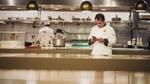 A chef consulting his phone in a professional kitchen while another chef works in the background
